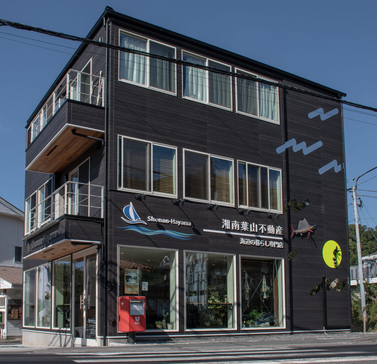 Modern dark-gray building with large white-framed windows and blue signage on a sunny day; red mailbox at street level outside the entrance.