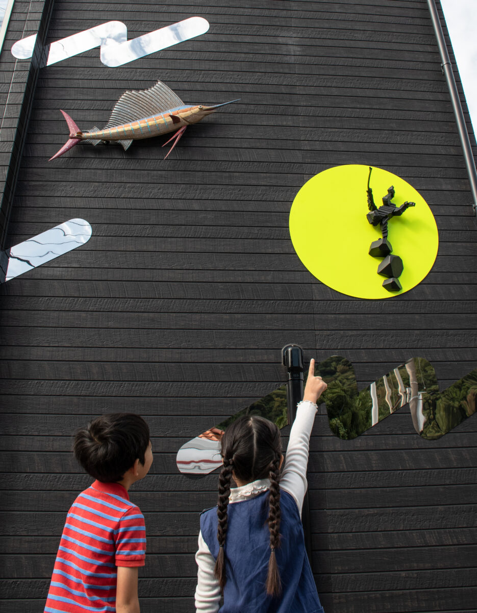 Children observe and point at decorative wall art on a black wooden wall, including a large fish sculpture and a bright yellow circle art piece.